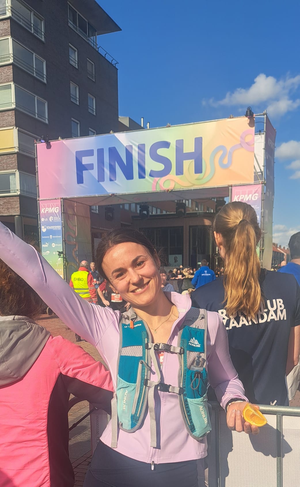 Konxhe smiling at the finish line of a road race, one arm raised and holding an orange slice